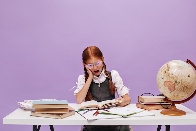 Shocked ginger girl with cool two pigtails in eyeglasses an school white and grey uniform reading book on purple background