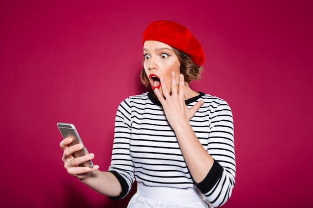 Free photo shocked ginger woman holding cheek while using smartphone over pink