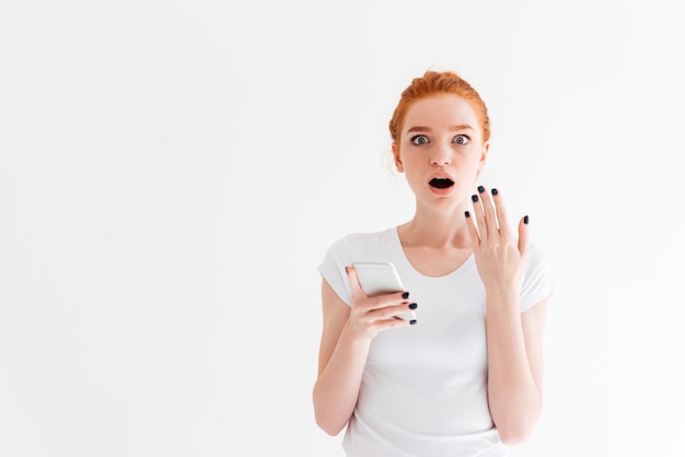 Shocked ginger woman in t-shirt holding smartphone and looking