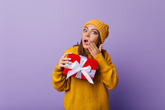 Shocked girl in casual sweater and hat posing with gift. Indoor portrait of debonair lady with new year present expressing amazement.