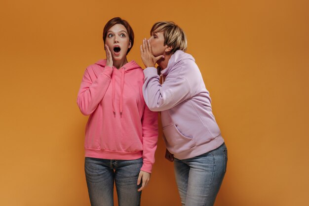 Shocked girl in jeans and wide pink hoodie listening to secret from her grandmother in stylish clothes on orange backdrop. 
