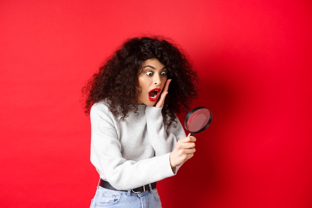 Shocked girl look through magnifying glass with dropped jaw seeing something amazing standing on red...