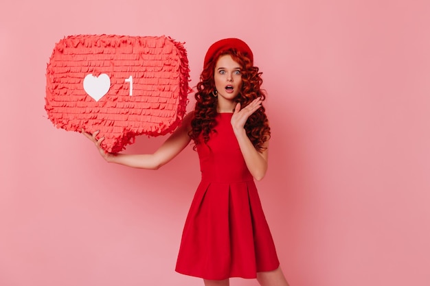 Free photo shocked girl looks at camera holding like sign on pink background portrait of woman in red dress and beret