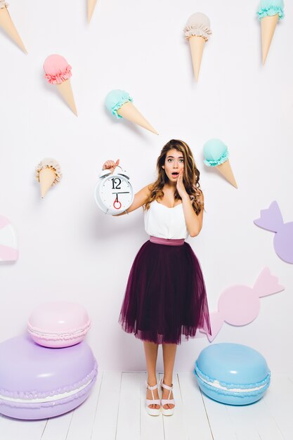 Shocked girl in midi skirt realized that guests were late for the her birthday party. Full-length portrait of trendy young woman with alarm clock and unhappy face expression posing near toy sweets.