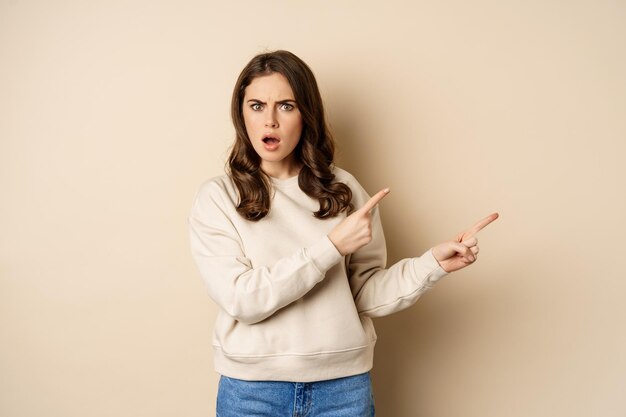 Shocked girl pointing fingers right gasping startled standing over beige background