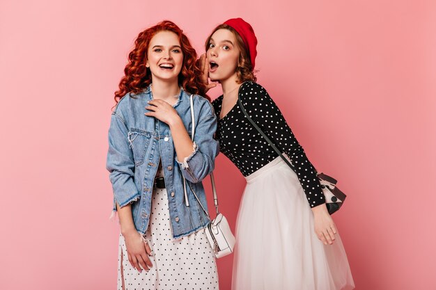 Shocked girl in red beret talking with friend. Attractive women talking on pink background.