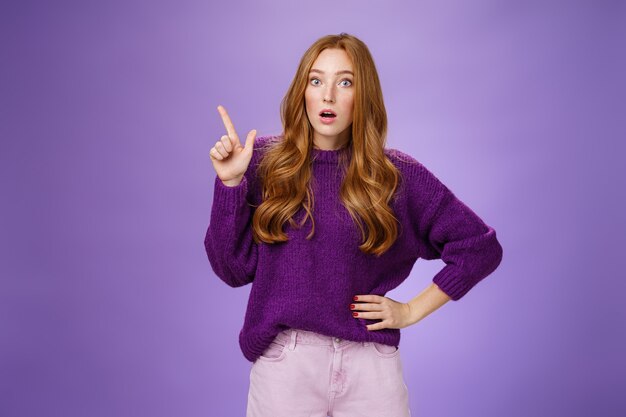 Shocked girl in stupor drop jaw and pop eyes at camera from amazement of unexpected stunning copy space pointing at upper left corner looking impressed and astonished over purpe background.