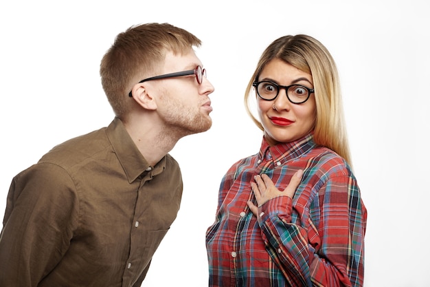 Shocked girl in styling checkered shirt and oval spectacles popping eyes out, holding hand on her chest, feeling terrified while some nerdy guy is going to kiss her, pouting his lips and closing eyes