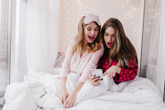 Shocked girl with long hairstyle looking at phone screen while sitting in bed. Portrait of chilling female friends in pyjamas posing in light bedroom.