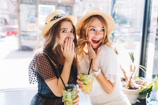 Shocked girls in hats holding glasses of cocktail looking away and emotionally covers faces