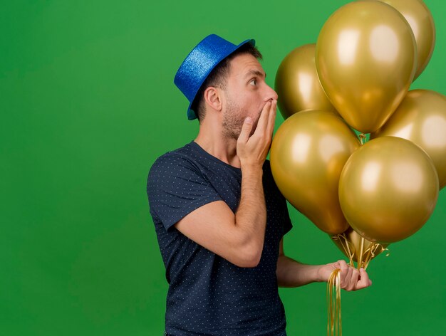 Shocked handsome caucasian man wearing blue party hat holds helium balloons puts hand on mouth isolated on green background with copy space