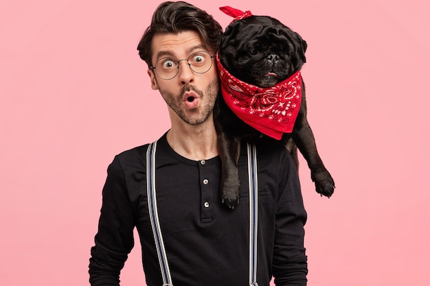 Shocked handsome male owner of dog, looks with terrible expression, finds out bad news, being in company with pet, dressed elegantly, pose together against pink wall. Emotions, lifestyle