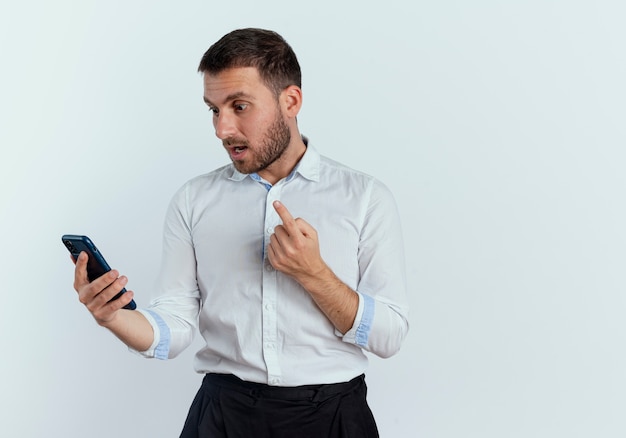 Shocked handsome man holds and looks at phone pointing up isolated on white wall