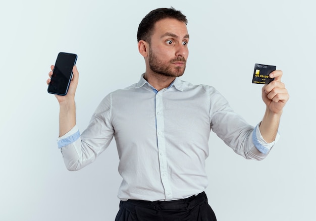 Шокированный красавец держит телефон и смотрит на кредитную карту, изолированную на белой стене