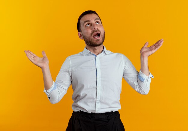 Shocked handsome man looks up with raised hands isolated on orange wall