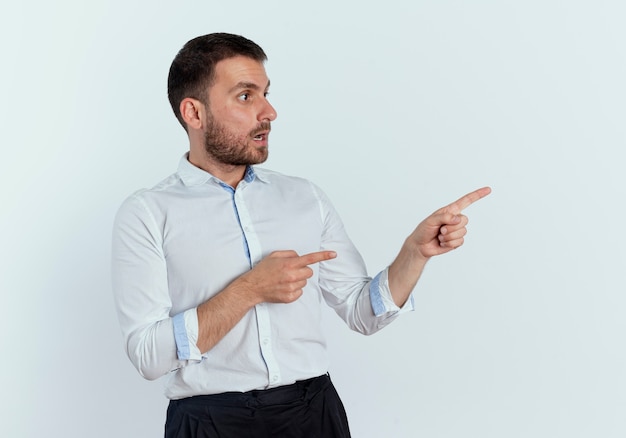 Shocked handsome man points and looks at side isolated on white wall