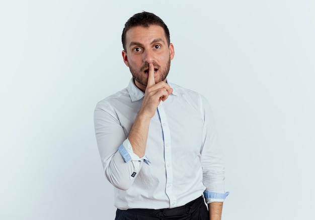 Shocked handsome man puts finger on mouth gesturing be quiet sign isolated on white wall