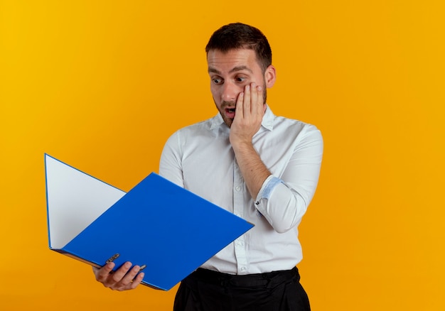 Shocked handsome man puts hand on face holding and looking at file folder isolated on orange wall