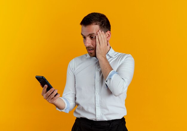 Shocked handsome man puts hand on face holding and looking at phone isolated on orange wall
