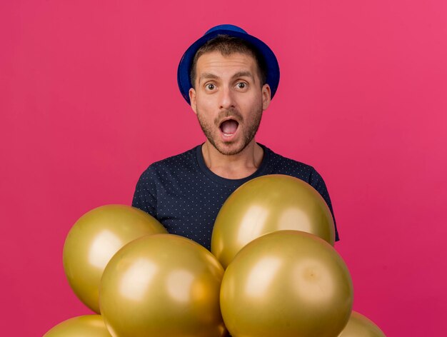 Shocked handsome man wearing blue party hat holds helium balloons isolated on pink wall with copy space