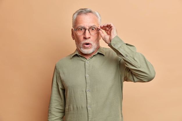 Shocked handsome stunned bearded elderly man has grey hair opens mouth widely keeps hand on spectacles cannot believe in shocking news wears formal shirt poses against brown studio wall