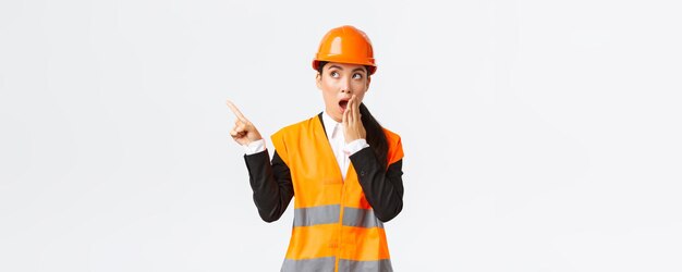 Shocked and impressed asian female engineer construction manager in safety helmet and reflective jacket gasping pointing finger upper left corner amazed found out something interesting