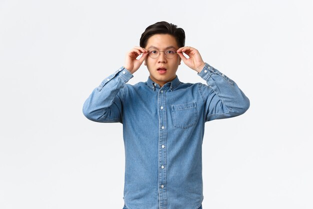 Shocked and impressed asian man put on glasses to see something, looking startled and amazed, witness big event, watching interesting announcement, standing white background