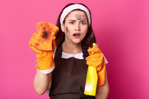 Shocked impressed cute young lady showing dirty washcloth, holding detergent in yellow bottle, opening mouth widely, wearing white handband, brown apron and casual t shirt. Household concept.