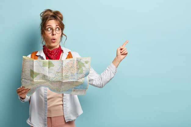 Shocked impressed female tourist points at copy space, holds travel map