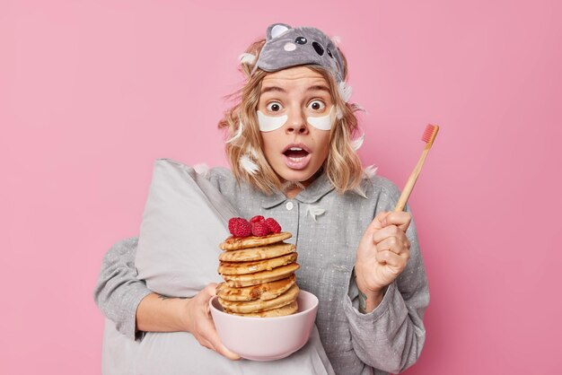 Shocked impressed young woman stares surprised holds bowl of pancakes and wooden toothbrush awakes early in morning applies beauty patches under eyes wears pajama sleepmask isolated over pink wall