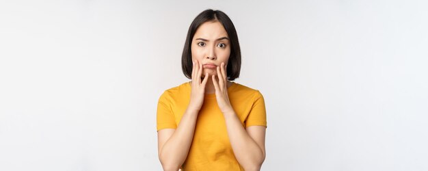 Shocked korean girl touching her face looking concerned at camera standing in yellow tshirt over white background