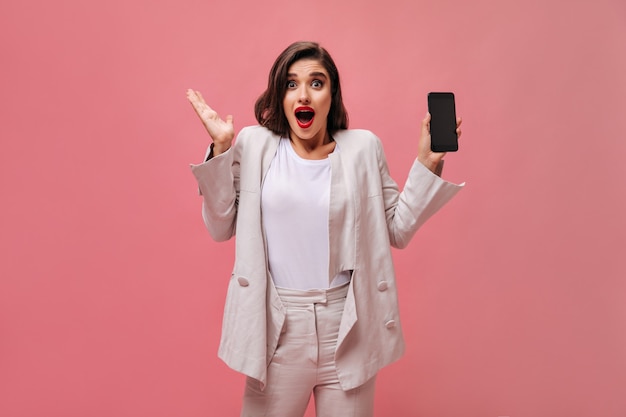 Shocked lady in cotton suit demonstrates phone on pink background.  Surprised woman with bright lips in white clothes holds phone in her hands.