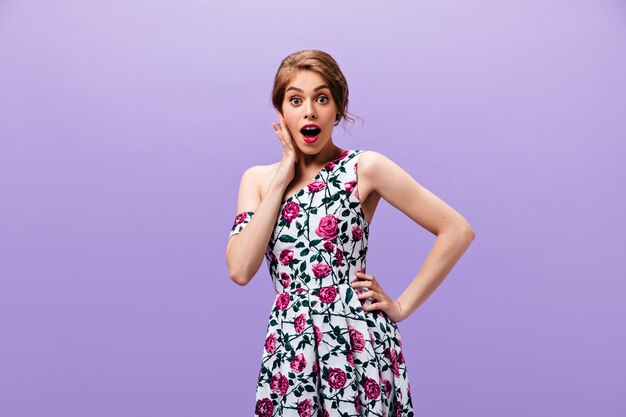 Shocked lady in floral dress poses on isolated background. Surprised young woman with red lips in stylish clothes looking into camera.