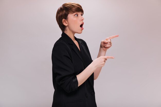 Shocked lady in suit points to place for text on white background. Surprised short-haired woman in black jacket poses on isolated backdrop