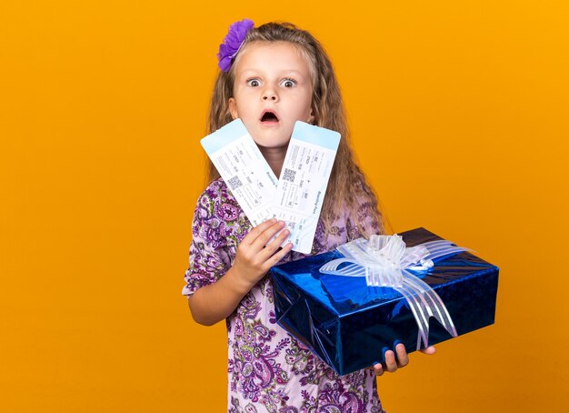 shocked little blonde girl holding gift box and air tickets isolated on orange wall with copy space