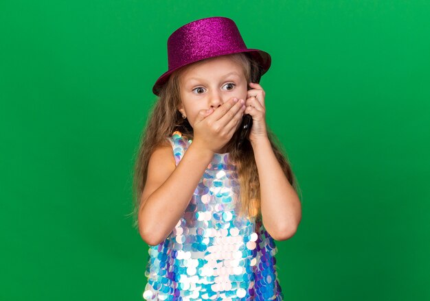 shocked little blonde girl with purple party hat putting hand on mouth talking on phone isolated on green wall with copy space