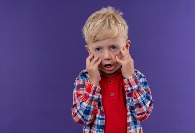 A shocked little boy with blonde hair wearing checked shirt holding hands on his face on a purple wall