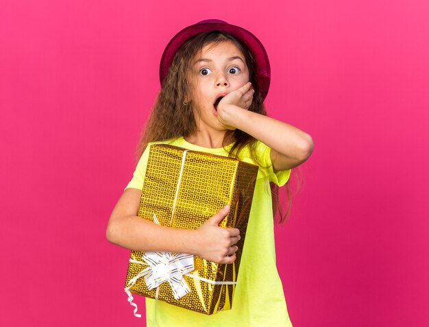 shocked little caucasian girl with purple party hat putting hand on mouth and holding gift box isolated on pink wall with copy space