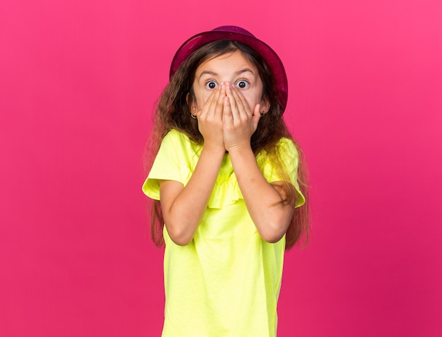 Free photo shocked little caucasian girl with purple party hat putting hands on mouth isolated on pink wall with copy space