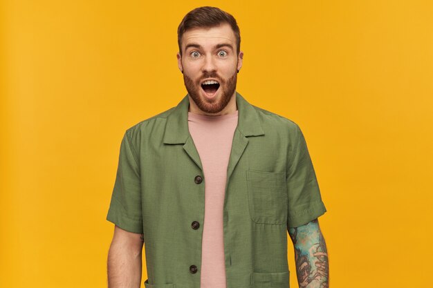 Shocked looking male, handsome guy with brunette hair and beard. wearing green short sleeves jacket. has tattoo and piercing. with open mouth, isolated over yellow wall