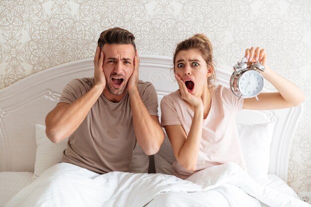 Shocked Lovely couple sitting together on bed with alarm clock