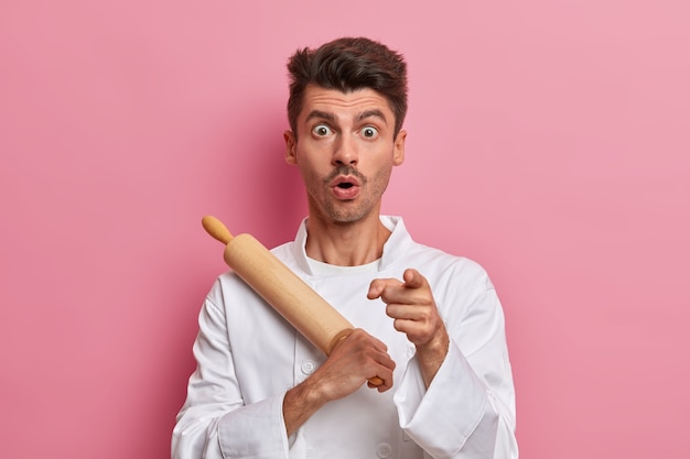 Shocked male baker holds rolling pin and points at front, gets ready to cook, works in kitchen, wears uniform