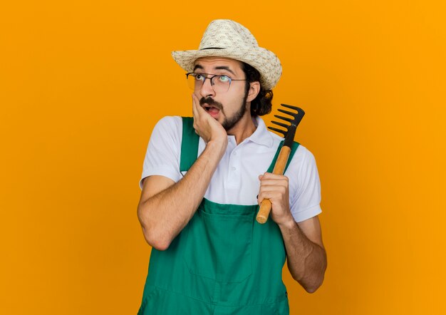 Shocked male gardener in optical glasses wearing gardening hat puts hand on chin and holds rake looking at side 