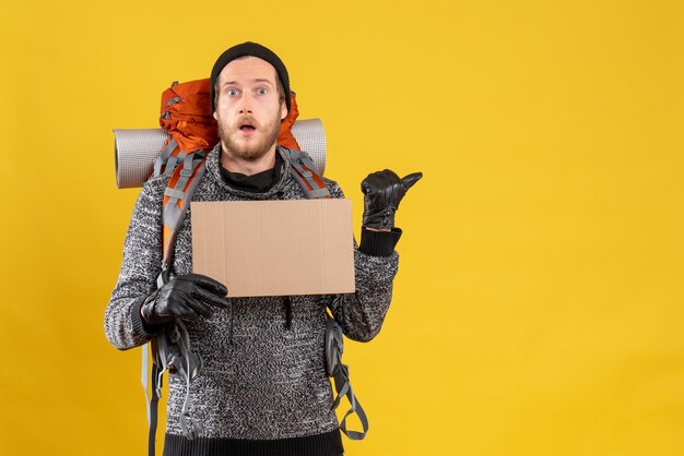 shocked male hitchhiker with leather gloves and backpack holding blank cardboard pointing at something