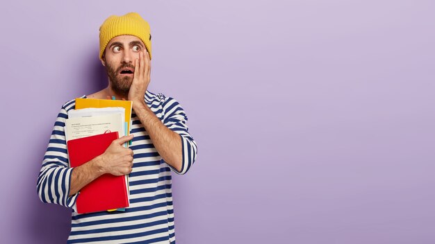 Shocked male student looks with terrified expression, keeps hand on cheek, carries notepad and papers, going to study, dressed in casual clothing stands indoor over violet wall