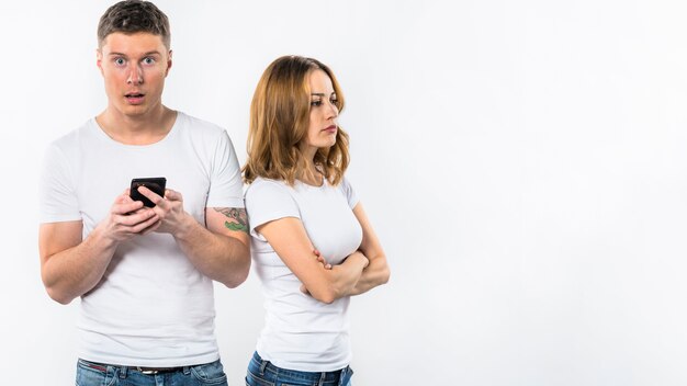 Шокированный мужчина держит мобильный телефон в руке, стоя возле ее злой подруги