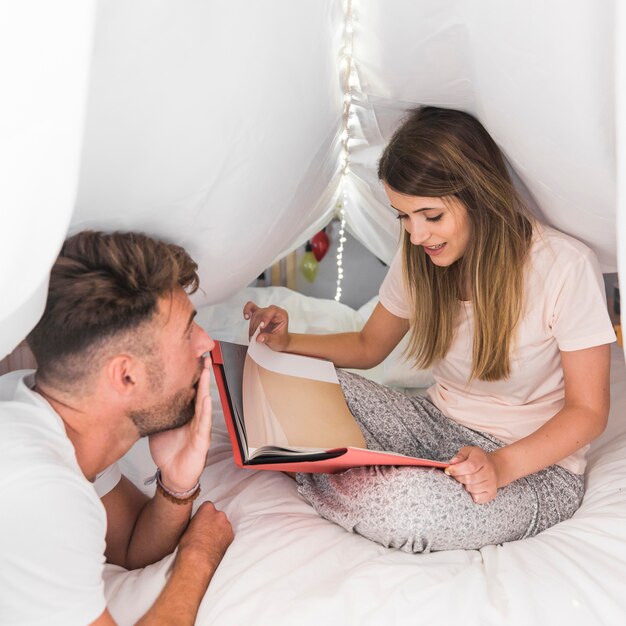 Shocked man looking at woman holding book sitting on bed under the curtain