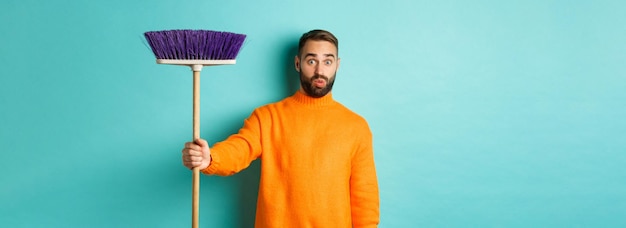 Shocked man receiving broom to do house chores looking confused standing over light blue background