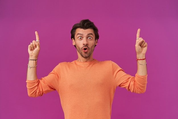 Shocked man with dark hair and bristle. wearing orange sweater with rolled up sleeves. has bracelets, rings