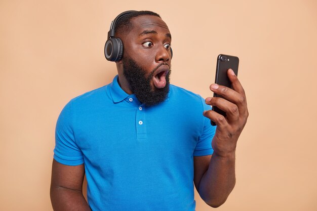 Shocked man with thick beard stares with unbelievable gaze at smartphone display wears wireless headphones on ears dressed in casual blue t shirt isolated over beige wall